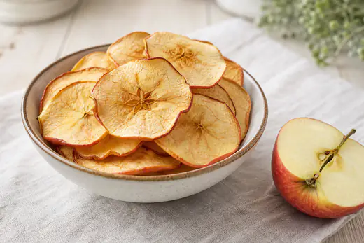 Dünne goldgelbe Apfelchips mit sichtbarem Kerngehäuse in einer Schale, daneben ein halber Apfel