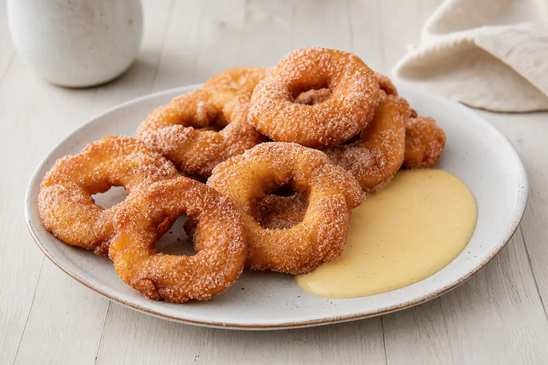 Goldbraune Apfelküchlein aus Apfelringen im Teig, mit Zimtzucker bestreut, auf einem Teller mit Vanillesoße