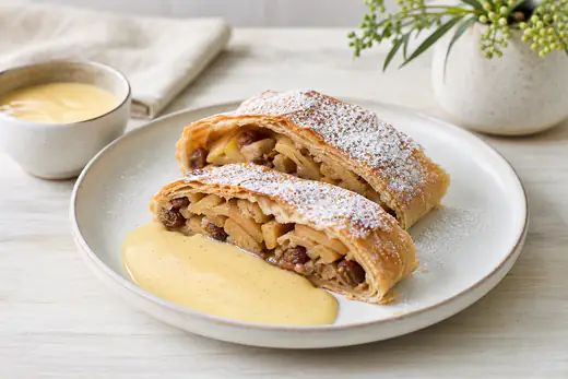 Goldbrauner Apfelstrudel, in Scheiben geschnitten, mit Puderzucker bestäubt und Vanillesoße auf einem Teller