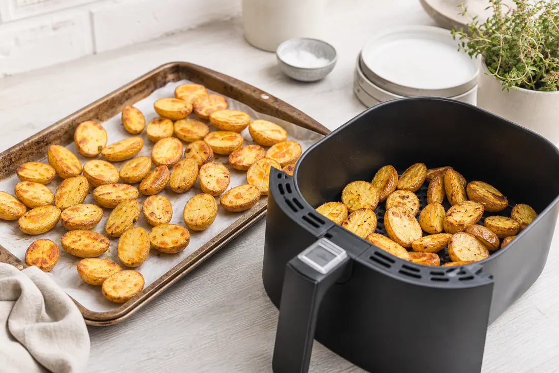 Backblech mit Ofenkartoffelhälften neben einem Airfryer-Korb mit den gleichen goldbraunen Kartoffeln