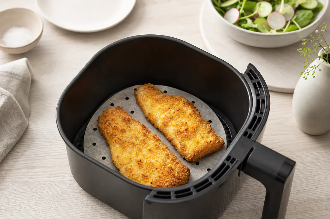 Airfryer-Korb mit einem zugeschnittenen, gelochten Backpapier und zwei goldbraunen Fischfilets darauf