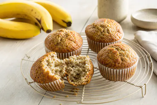 Vier goldbraune Bananenmuffins in Papierförmchen auf einem Abkühlgitter, einer aufgebrochen, dahinter reife Bananen