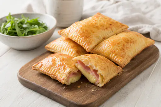Goldbraune rechteckige Blätterteigtaschen mit Schinken-Käse-Füllung, eine aufgeschnitten, auf einem Holzbrett