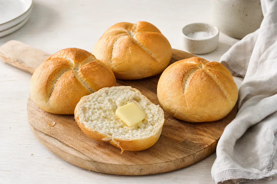 Vier frisch aufgebackene goldbraune Brötchen mit knuspriger Kruste auf einem Brett, eines aufgeschnitten mit Butter