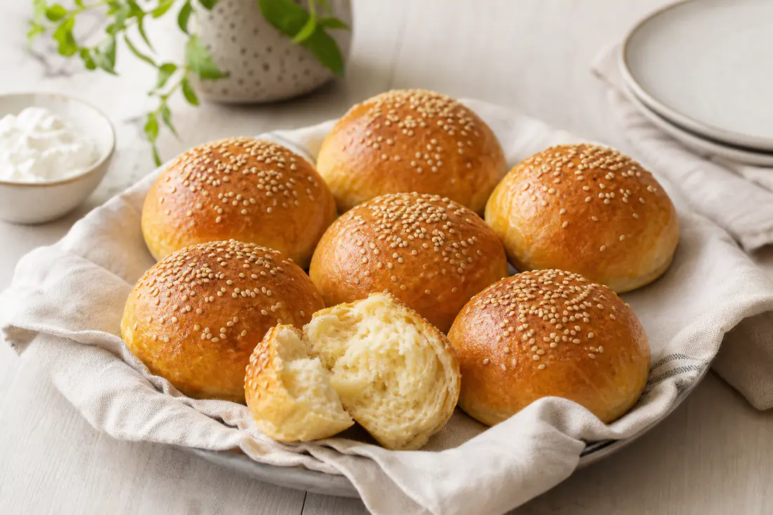 Sechs selbst gebackene Quarkbrötchen mit Sesam und goldbrauner Kruste auf einem Küchentuch, eines aufgebrochen
