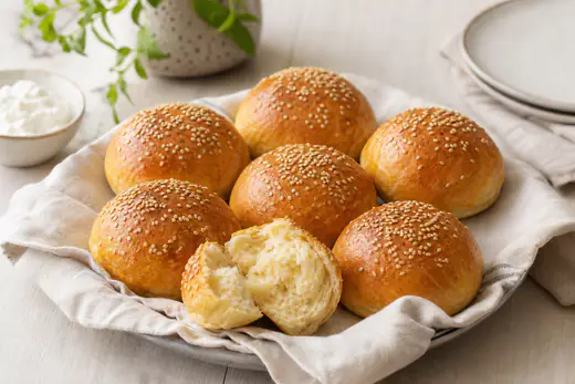 Sechs selbst gebackene Quarkbrötchen mit Sesam und goldbrauner Kruste auf einem Küchentuch, eines aufgebrochen