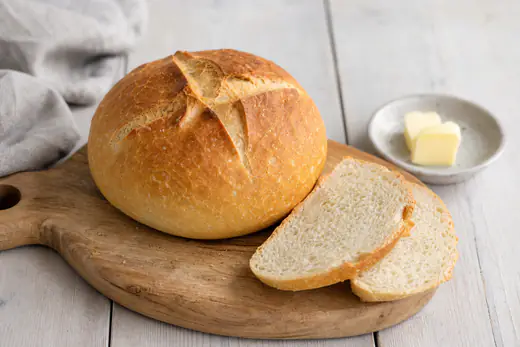 Kleines rundes Weißbrot mit goldbrauner Kruste und kreuzförmigem Einschnitt auf einem Holzbrett, mit zwei Scheiben und einer Butterdose