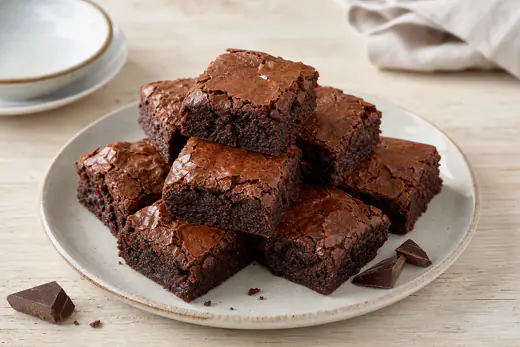 Neun quadratische Schokoladen-Brownies mit rissiger glänzender Oberfläche, gestapelt auf einem Teller mit Schokostückchen