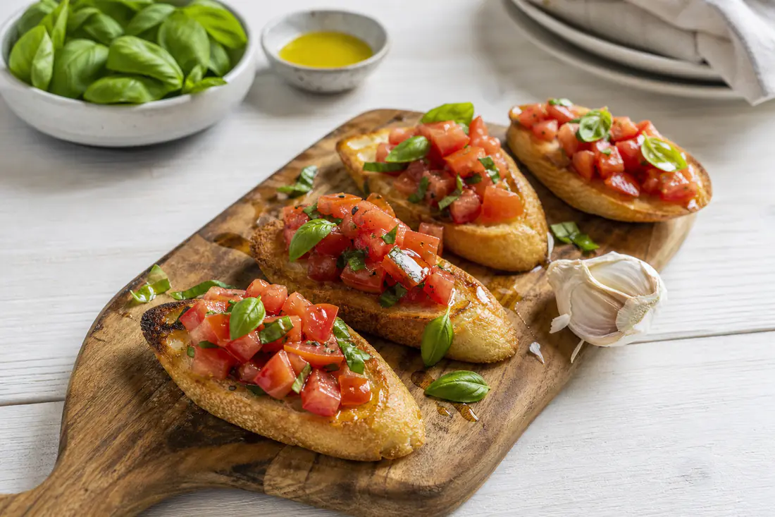 Geröstete Baguettescheiben mit gewürfelten Tomaten, Basilikum und Olivenöl auf einem Holzbrett, daneben eine halbierte Knoblauchzehe
