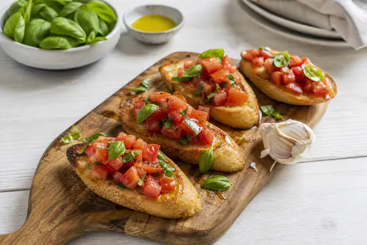 Geröstete Baguettescheiben mit gewürfelten Tomaten, Basilikum und Olivenöl auf einem Holzbrett, daneben eine halbierte Knoblauchzehe