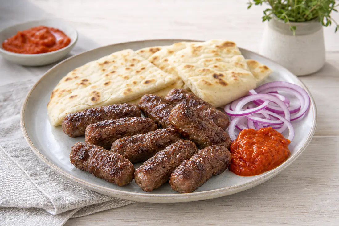 Acht fingerförmige gegrillte Cevapcici auf einem Teller mit Fladenbrot, Ajvar und rohen Zwiebelringen
