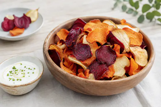 Holzschüssel mit bunten dünnen Gemüsechips aus Rote Bete, Möhre und Pastinake, daneben ein Schälchen Joghurt-Dip