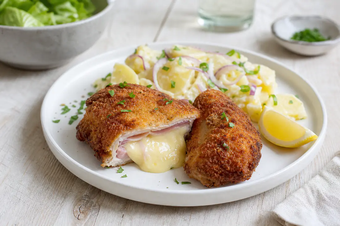 Ein aufgeschnittenes goldbraun paniertes Cordon bleu mit Schinken- und Käsefüllung, dazu Kartoffelsalat und eine Zitronenspalte