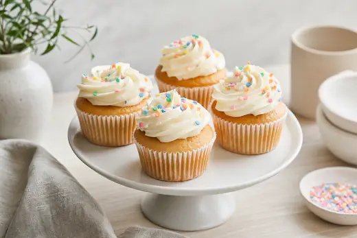 Vier Vanille-Cupcakes in Papierförmchen mit weißen Frischkäse-Frosting-Häubchen und bunten Streuseln auf einer weißen Etagere