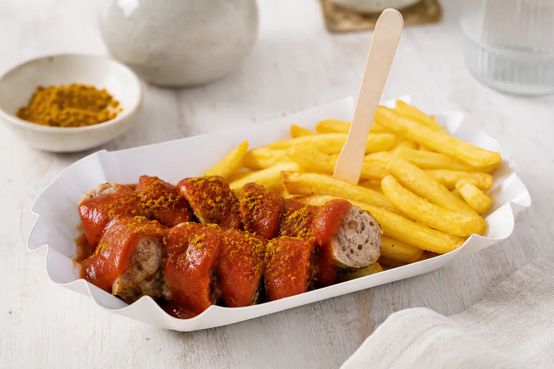 Eine in Scheiben geschnittene Bratwurst mit roter Currysoße und Currypulver auf einem Pappteller, dazu goldene Pommes und eine Holzgabel