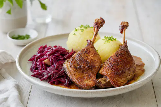 Zwei Entenkeulen mit knuspriger goldbrauner Haut auf einem Teller, dazu Rotkohl und Kartoffelklöße