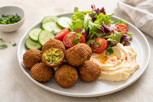 Runde knusprige Falafel, eine aufgebrochen mit grüner Kräuterfüllung, auf einem Teller mit Hummus, Gurkenscheiben, Tomaten und Salat