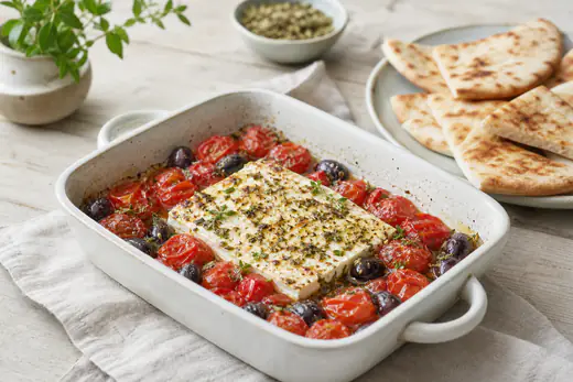 Gebackener Feta mit geplatzten Kirschtomaten, Oliven und Oregano in einer kleinen ofenfesten Form, daneben Brot