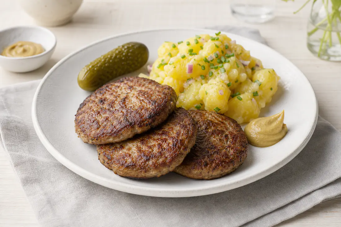 Drei flache goldbraune Frikadellen auf einem weißen Teller mit Kartoffelsalat, Senf und einer Gewürzgurke