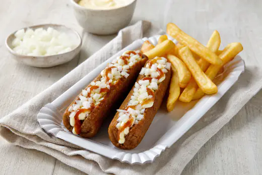 Zwei goldbraune Frikandeln, längs eingeschnitten und mit Mayonnaise, Currysauce und gehackten Zwiebeln belegt, dazu Pommes auf einem Pappteller