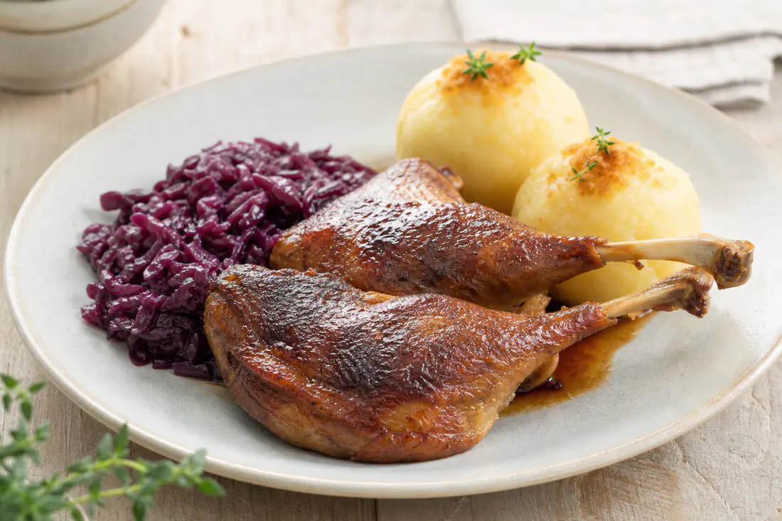 Zwei Gänsekeulen mit knuspriger dunkelbrauner Haut auf einem Teller, dazu Rotkohl und Kartoffelklöße