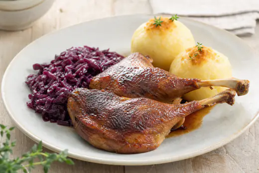 Zwei Gänsekeulen mit knuspriger dunkelbrauner Haut auf einem Teller, dazu Rotkohl und Kartoffelklöße