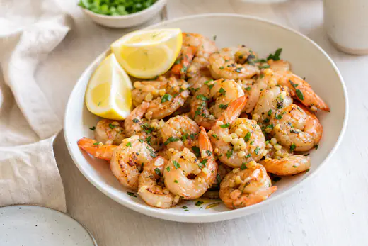 Gegarte rosa Garnelen mit Knoblauch, Petersilie und Zitronenspalten in einer flachen Schale, an den Rändern leicht gebräunt