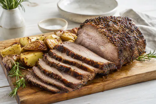 Aufgeschnittener Schweinenacken-Braten mit dunkler Gewürzkruste auf einem Holzbrett, daneben Ofenkartoffeln und Rosmarin