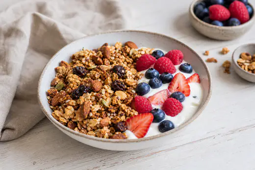 Goldbraune Granola-Cluster mit Nüssen und Rosinen in einer Schale, daneben Joghurt und frische Beeren
