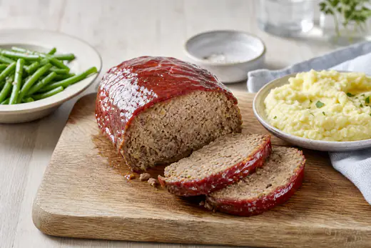 Aufgeschnittener Hackbraten mit glänzender Ketchup-Glasur auf einem Holzbrett, dazu Kartoffelpüree und grüne Bohnen