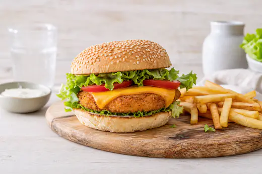 Hähnchenburger mit knusprigem Patty, Käse, Salat und Tomate im Sesambrötchen auf einem Holzbrett, daneben Pommes