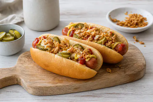 Zwei Hot Dogs in weichen Brötchen mit Würstchen, Ketchup, Senf, Remoulade, Röstzwiebeln und Gewürzgurkenscheiben auf einem Holzbrett