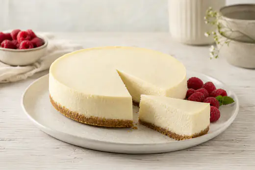 Runder Käsekuchen mit Keksboden und heller cremiger Füllung, ein Stück auf dem Kuchenteller, daneben frische Himbeeren