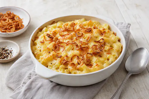 Kleine runde Form mit überbackenen Käsespätzle, goldener Käsekruste und knusprigen Röstzwiebeln, daneben ein Löffel