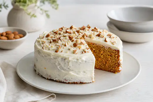 Runder Karottenkuchen mit Frischkäse-Topping und gehackten Mandeln, ein Stück angeschnitten, auf einem Kuchenteller