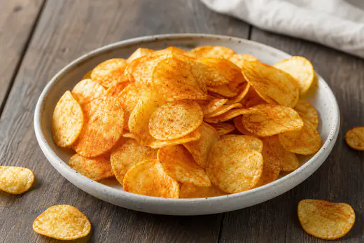 Schüssel mit dünnen runden goldgelben Kartoffelchips mit Paprikagewürz, dazu einzelne Chips auf einem dunklen Holztisch