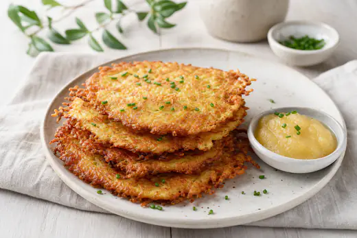 Mehrere goldbraune knusprige Kartoffelpuffer auf einem Teller, daneben eine Schale Apfelmus und etwas Schnittlauch