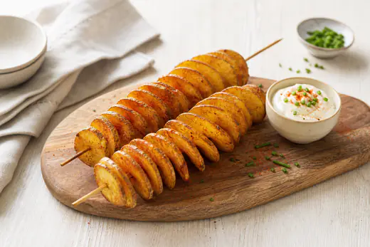 Zwei knusprige goldbraune Kartoffelspiralen auf Holzspießen mit Paprikagewürz, auf einem Holzbrett mit einem Schälchen Dip