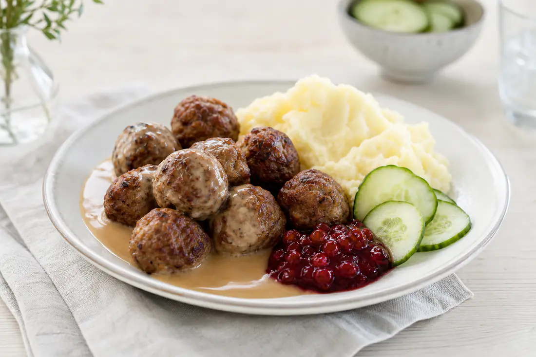 Goldbraune runde Köttbullar auf einem Teller mit Kartoffelpüree, Preiselbeeren, Rahmsoße und einer Gurkenscheibe