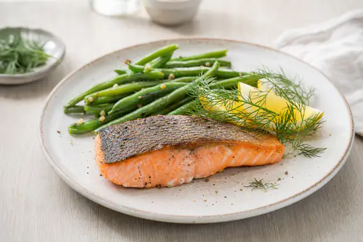 Gegartes Lachsfilet mit knuspriger Haut auf einem Teller, dazu Zitronenspalten, frischer Dill und grüne Bohnen