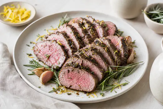 Aufgeschnittenes rosa gebratenes Lammfilet mit Kräuterkruste, fächerförmig auf einem Teller mit Rosmarin, Knoblauch und Zitronenabrieb