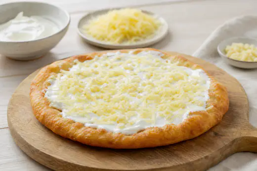 Goldbraunes rundes Langos-Fladenbrot mit Schmand, geriebenem Käse und Knoblauch auf einem Holzbrett