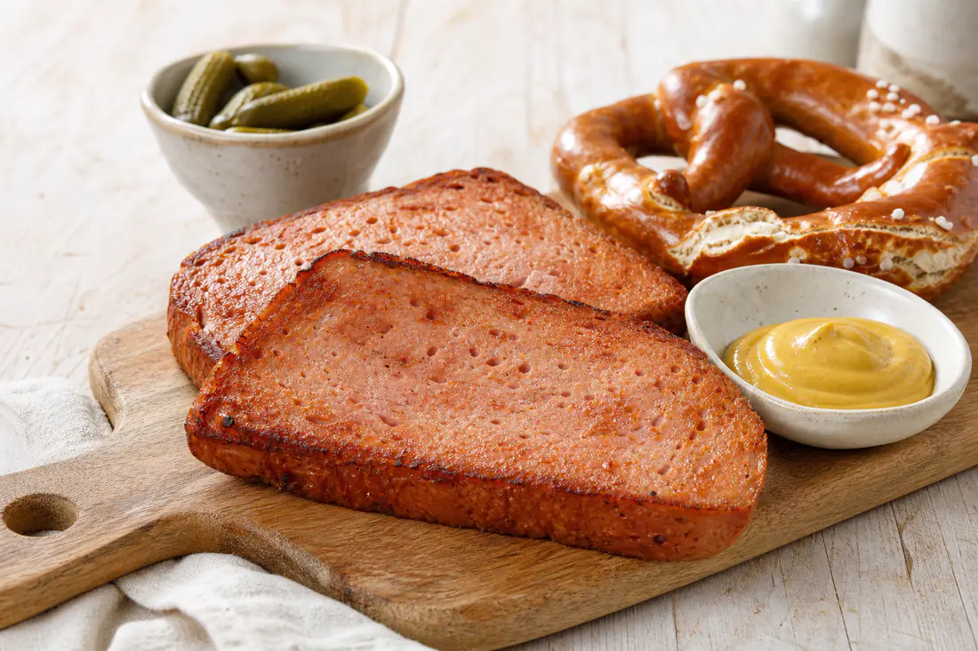 Zwei dicke, goldbraun gebackene Leberkäse-Scheiben mit knuspriger Kruste, dazu süßer Senf und eine Laugenbrezel auf einem Holzbrett