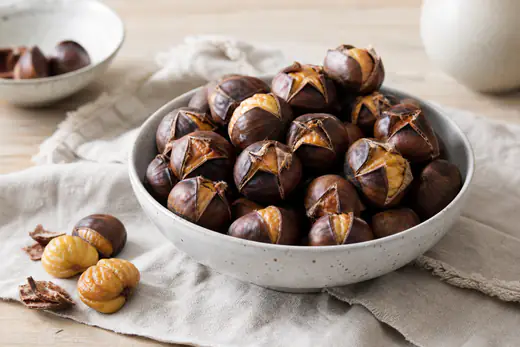 Schüssel mit gerösteten Maronen mit kreuzförmig eingeschnittener, aufgeplatzter Schale, daneben geschälte Maronen auf einem Holztisch