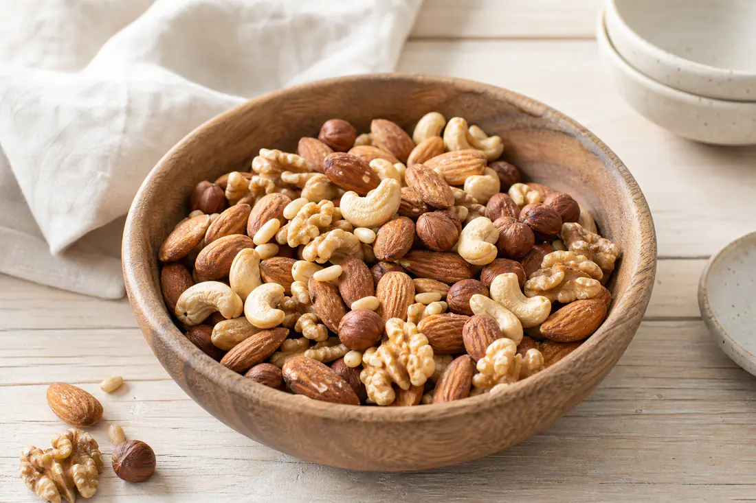 Geröstete Mandeln, Walnüsse, Haselnüsse und Cashews in einer Holzschale, daneben ein paar Pinienkerne