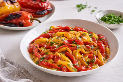 Geröstete rote und gelbe Paprikastreifen ohne Haut mit Olivenöl, Knoblauch und Petersilie in einer flachen Schale, daneben ganze Paprika