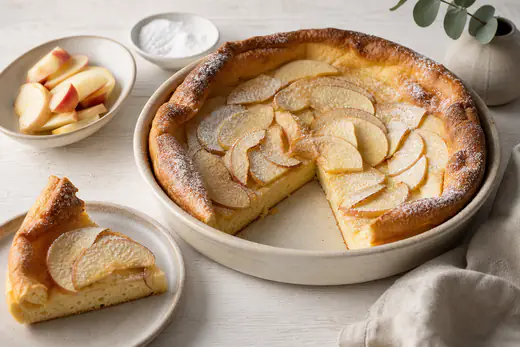 Aufgegangener goldbrauner Ofenpfannkuchen in runder Form mit dünnen Apfelscheiben und Puderzucker, ein Stück auf einem Teller
