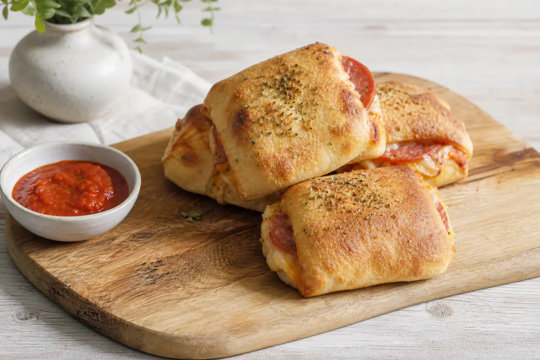 Goldbraune gefüllte Pizzabrötchen mit herausquellendem Mozzarella und Salami auf einem Brett, daneben eine Schale Tomatensauce