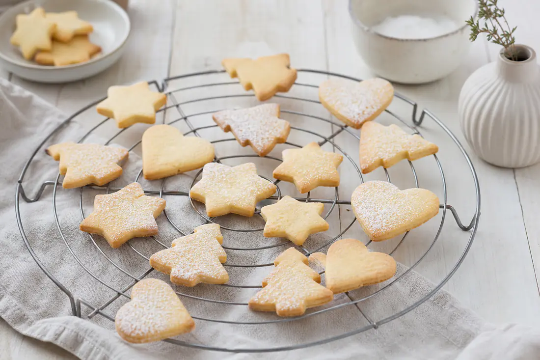 Goldgelbe Butterplätzchen in Sternform, Herzform und Tannenbaumform auf einem Kuchengitter, teils mit Puderzucker bestäubt