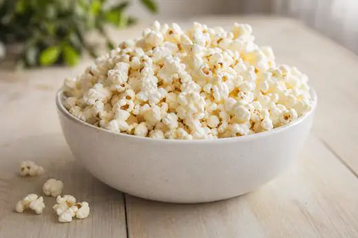 Frisch gepoppter, leicht gesalzener Popcorn in einer weißen Schüssel auf einem Holztisch, daneben einzelne Maiskörner